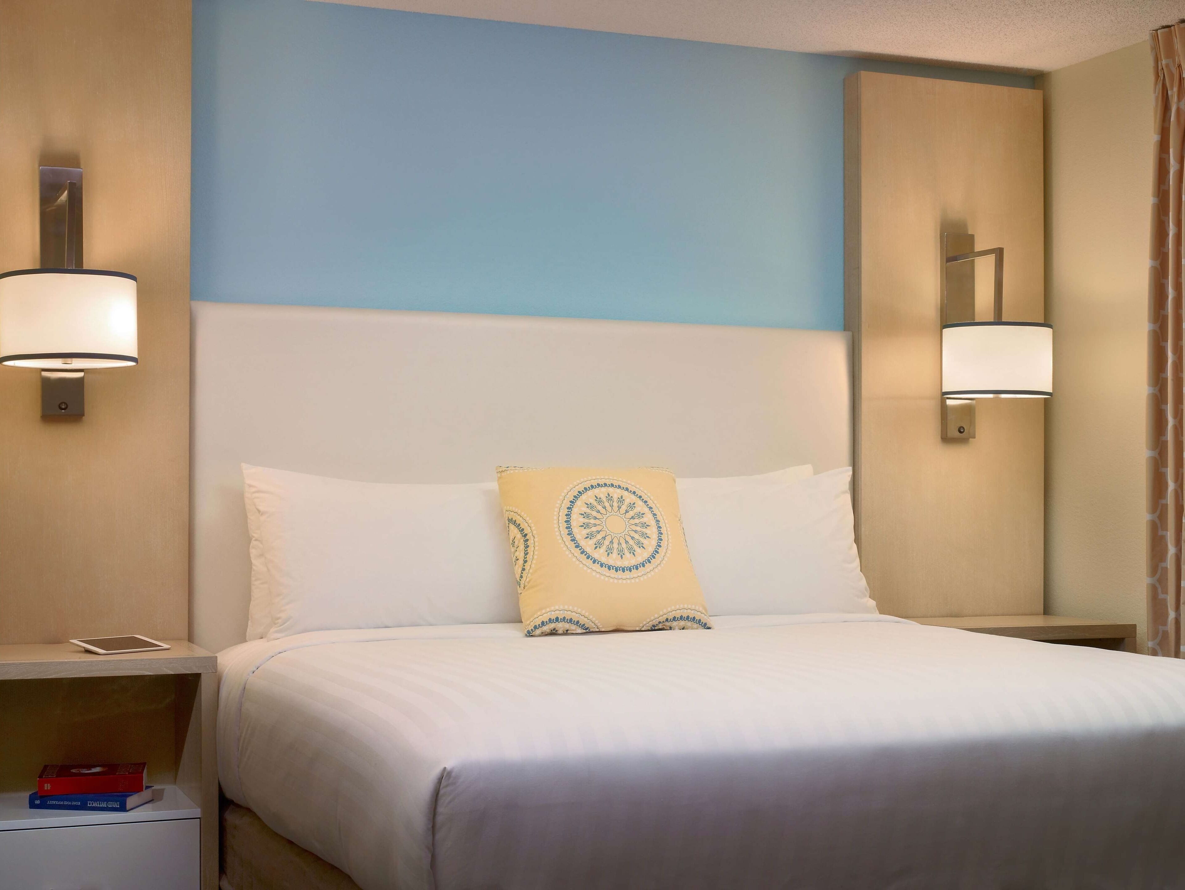 A hotel room with a bed, two bedside lamps, a nightstand with a tablet and books, and a blue wall.