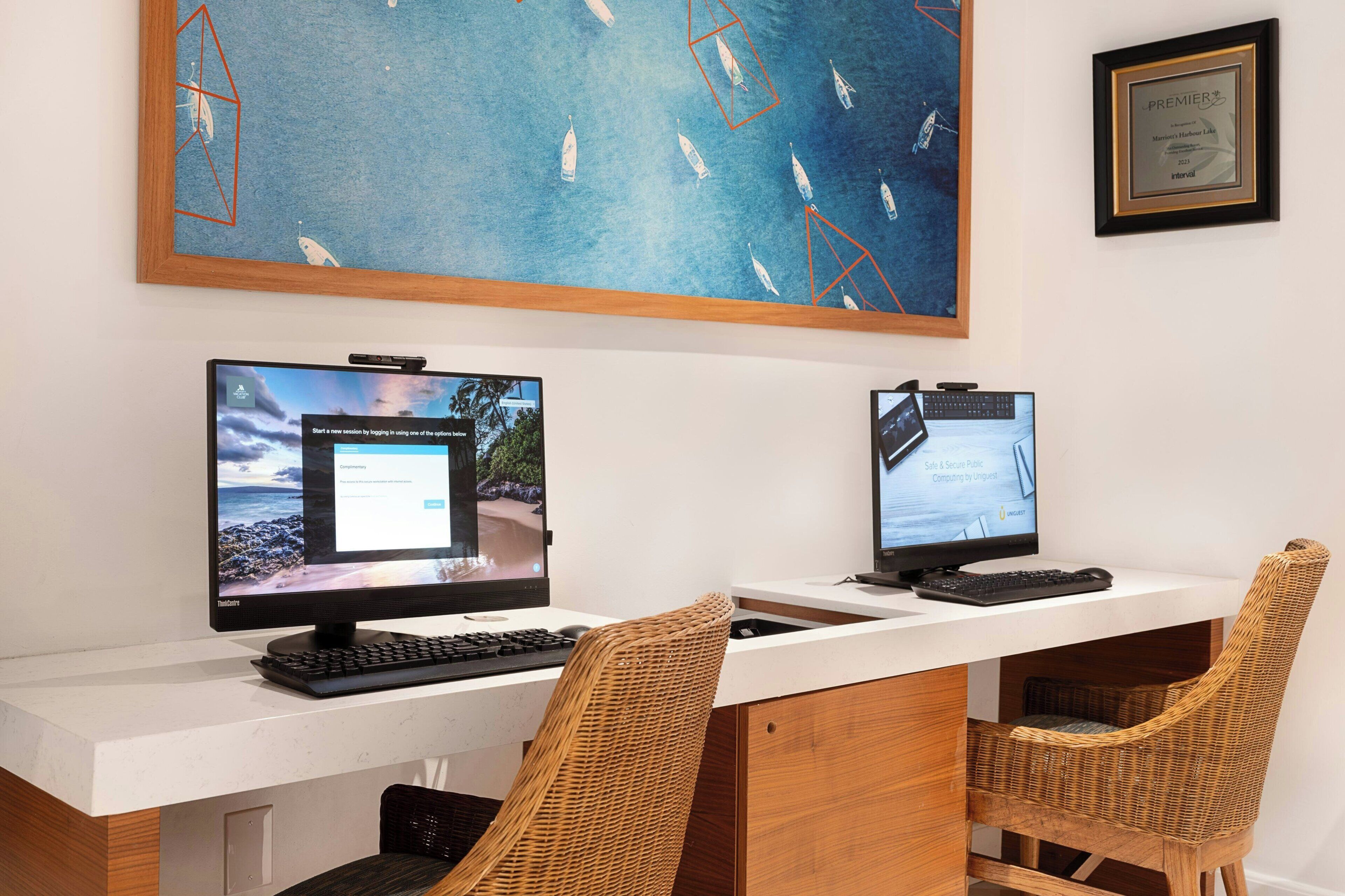 A workspace with two monitors, a keyboard, and a mouse on a white desk. There are two wicker chairs. Above the desk, there is a framed certificate and a large painting of boats on water.