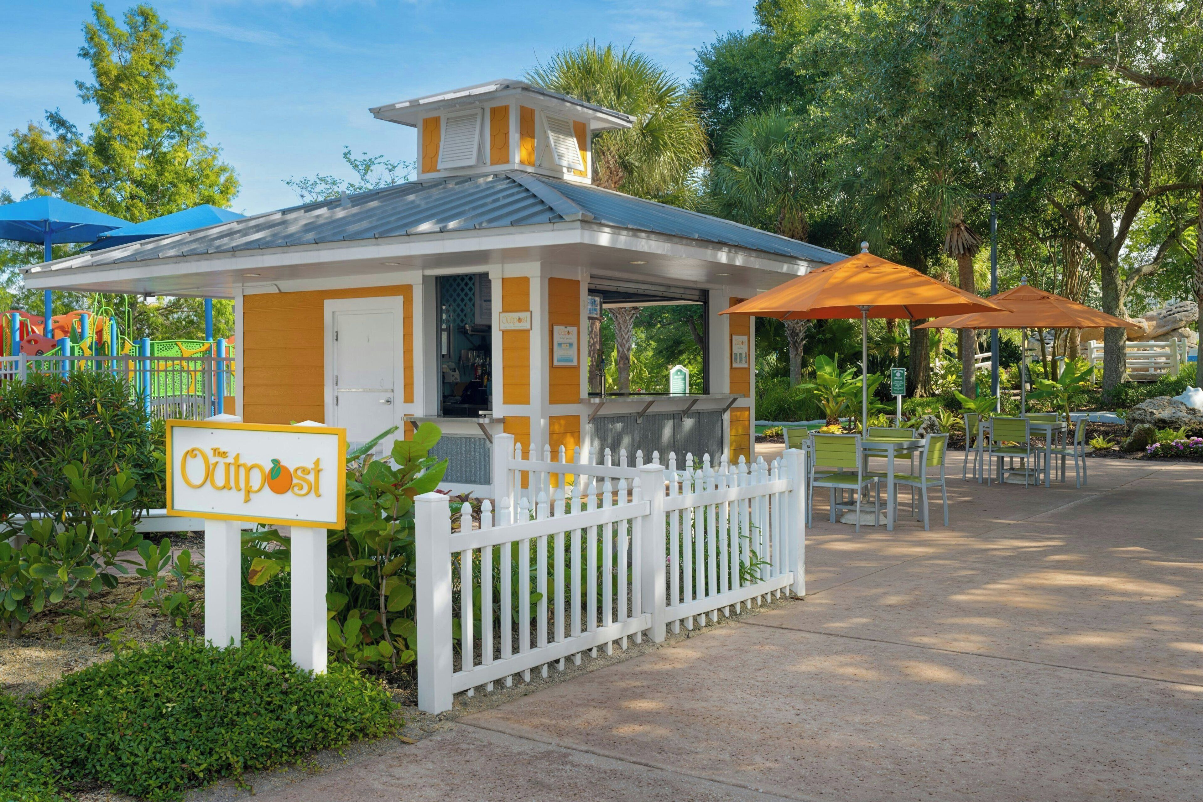 Un petit café en plein air, avec une clôture blanche en bois, une enseigne indiquant «The Outpost» et des tables à l’extérieur protégées par des parasols.
