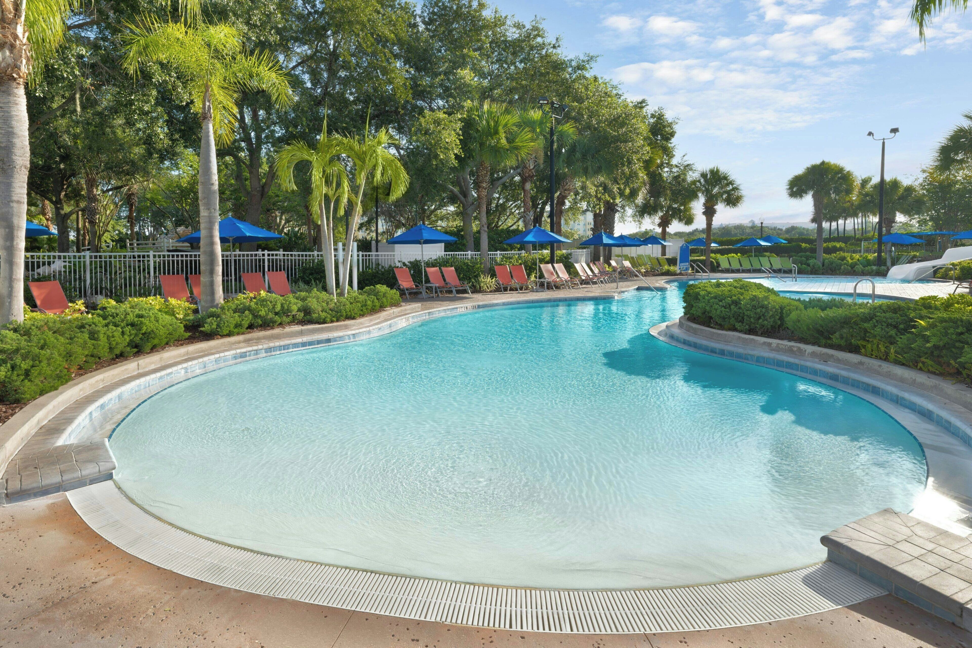 A clear swimming pool with a tiled edge, surrounded by a white fence, and shaded by blue umbrellas.