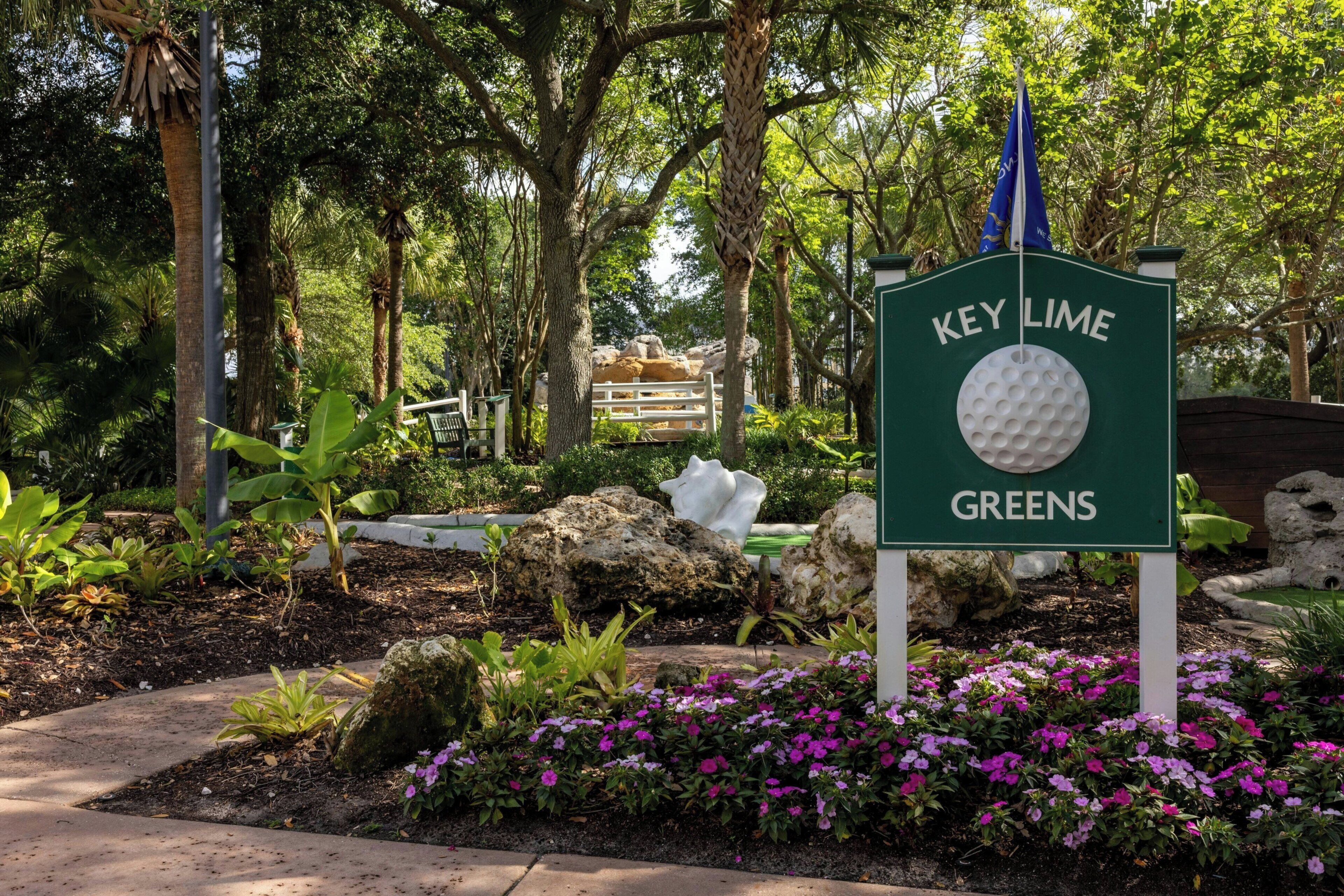 A sign for Key Lime Greens with a golf ball symbol, surrounded by lush greenery and flowering plants.