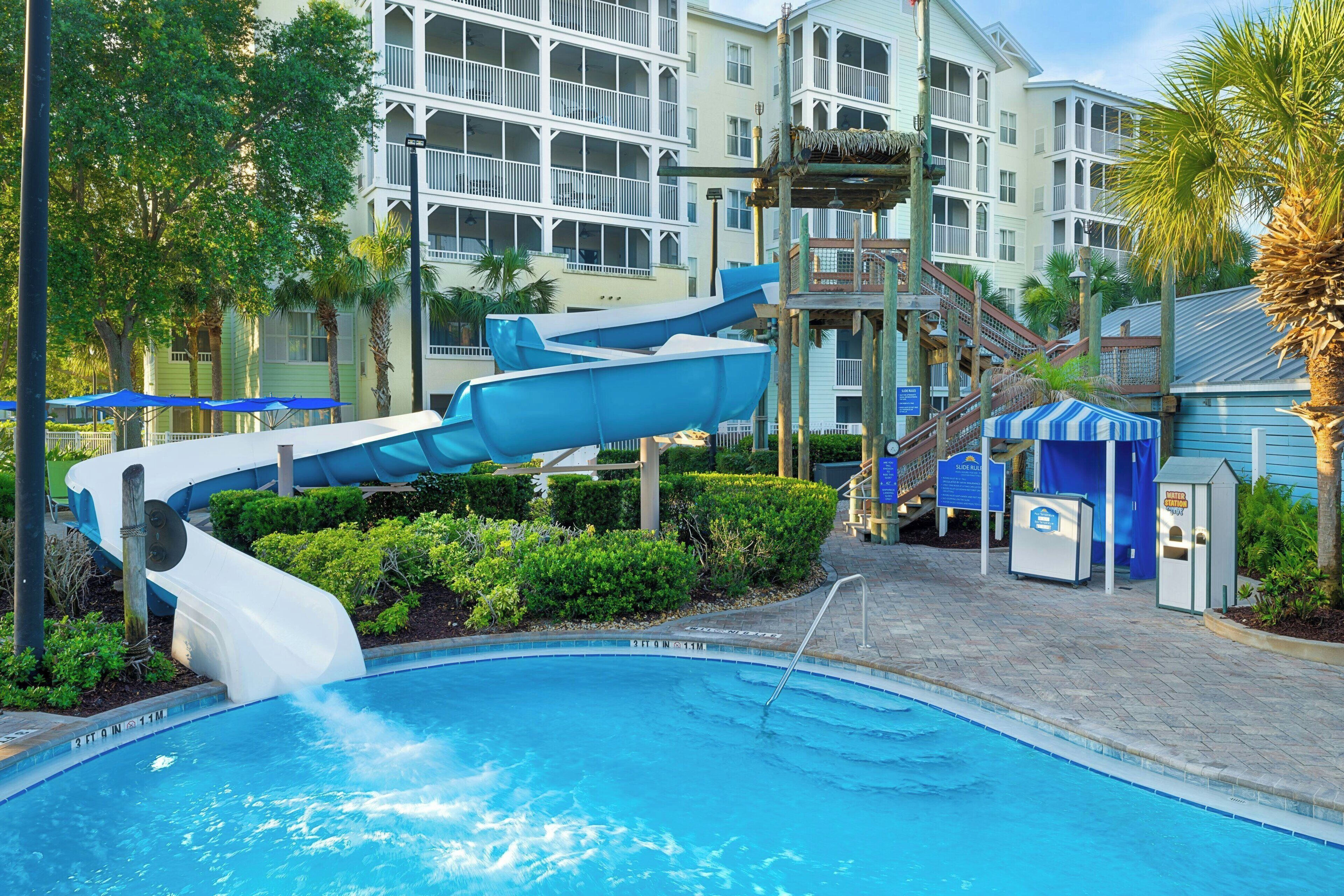 A pool with a water slide, a water station, and a building with balconies in the background.