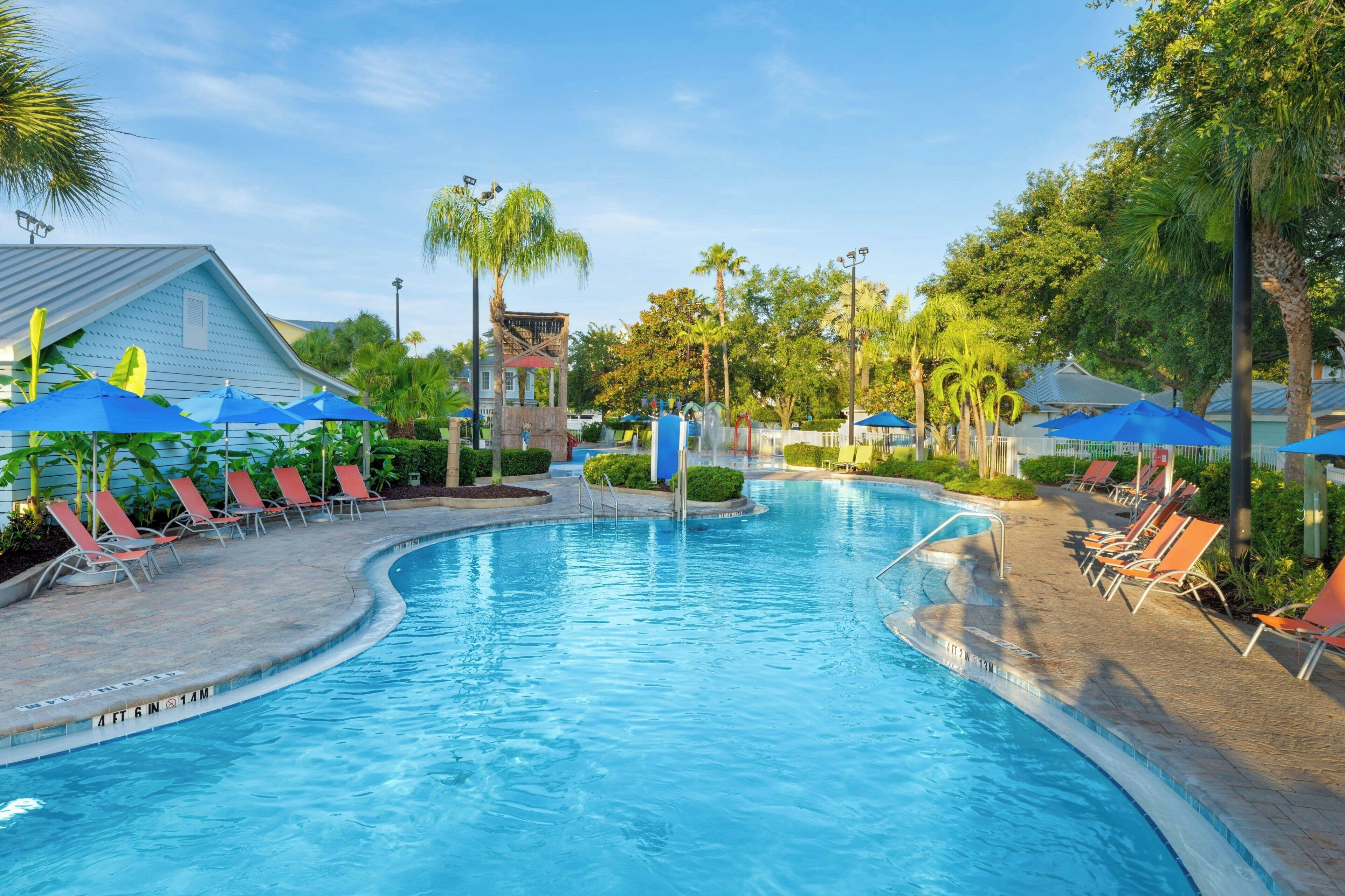 Ein Swimmingpool mit Liegestühlen und Sonnenschirmen, umgeben von Palmen und einem Gebäude mit geneigtem Dach.