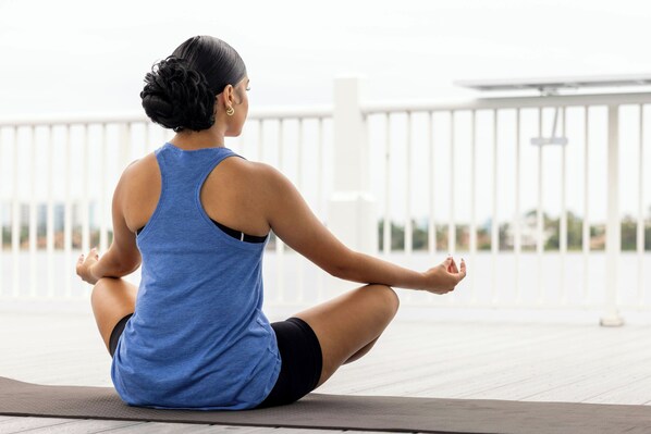 A person sitting on a yoga mat outdoors with a railing and a building in the background.