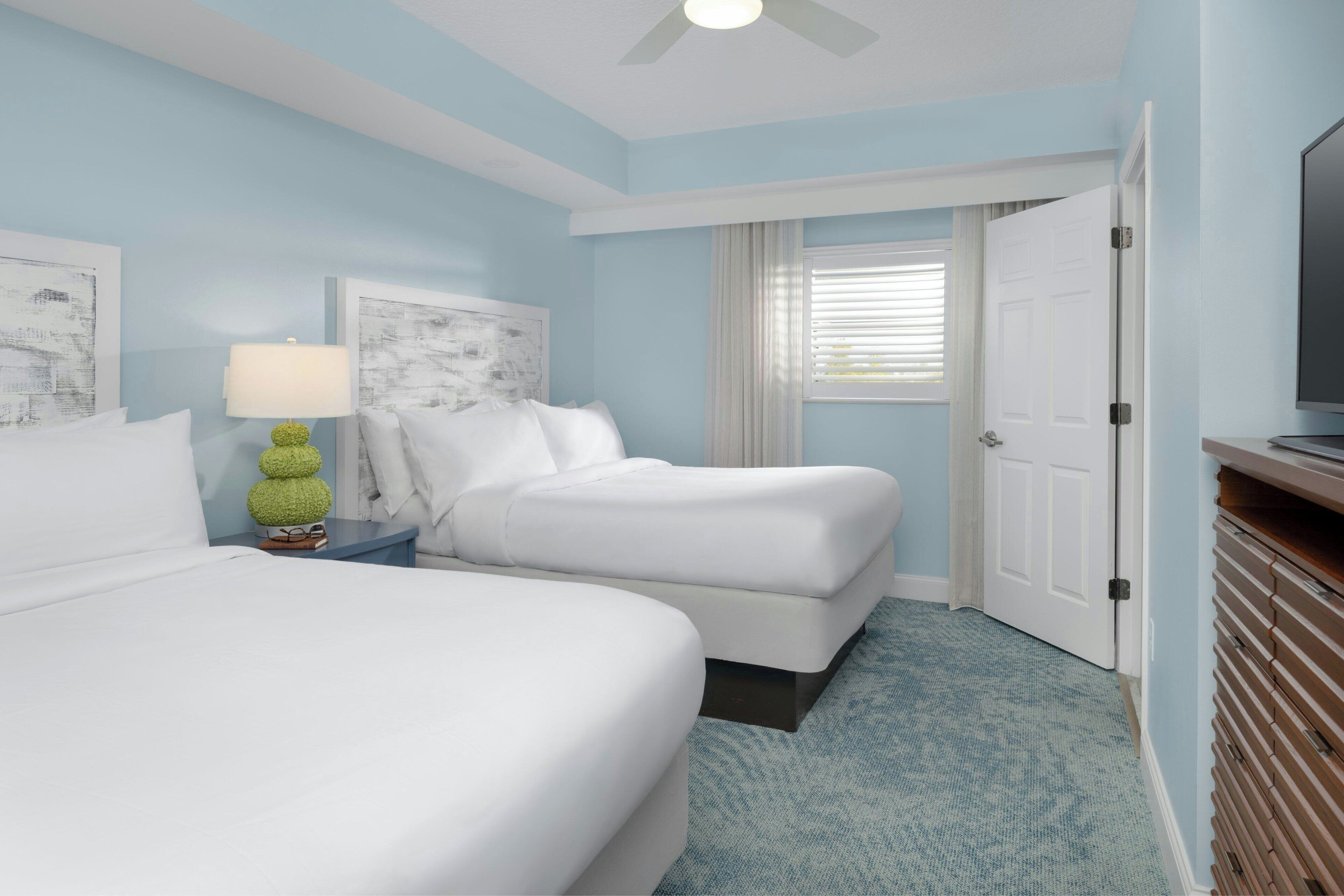 A hotel room with two beds, a nightstand with a lamp, a television, and a door leading to another room.