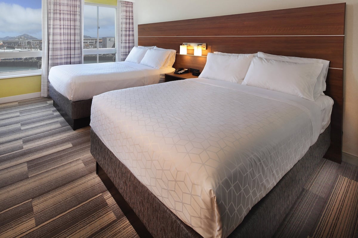 A hotel room with two beds, a large window with curtains, a wooden headboard, and a nightstand with a lamp.