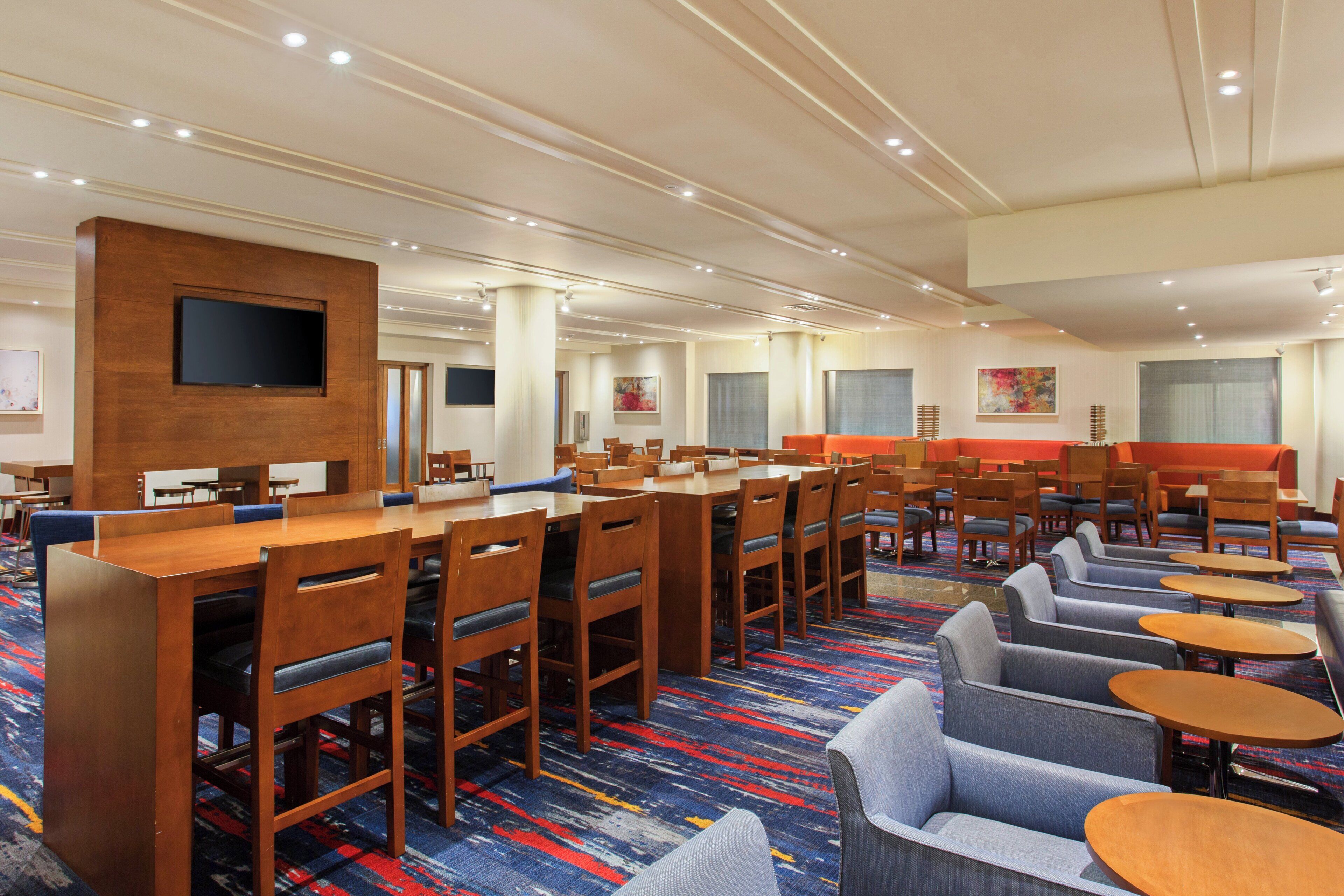 A hotel lobby with multiple seating areas, including tables and chairs, a flat-screen TV mounted on a wooden panel, and artwork on the walls.