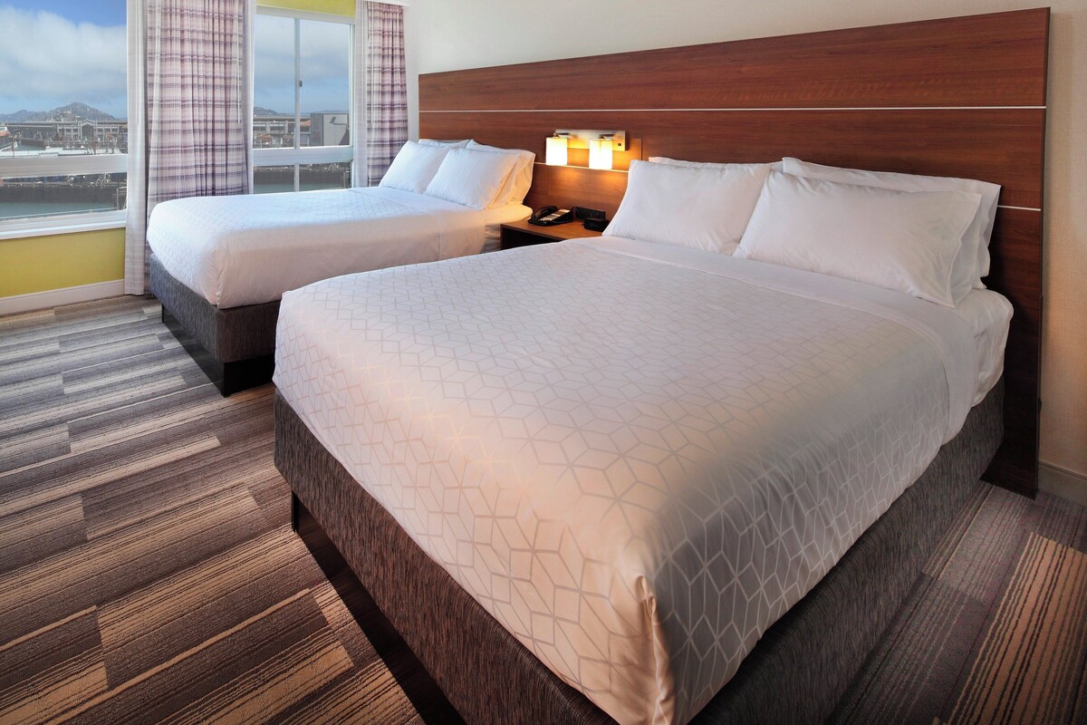 A hotel room with two beds, a wooden headboard, a nightstand with a lamp, and a large window with curtains.