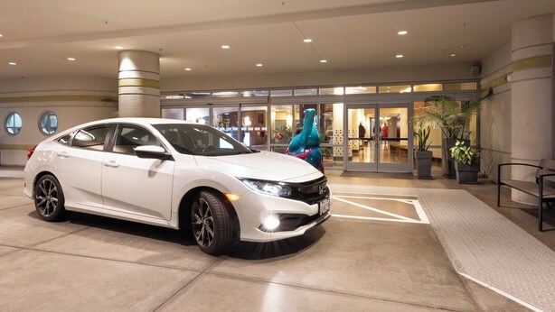 Une voiture blanche est garée dans un espace intérieur moderne, doté d’un plafond circulaire, de grandes fenêtres et de plantes en pot.