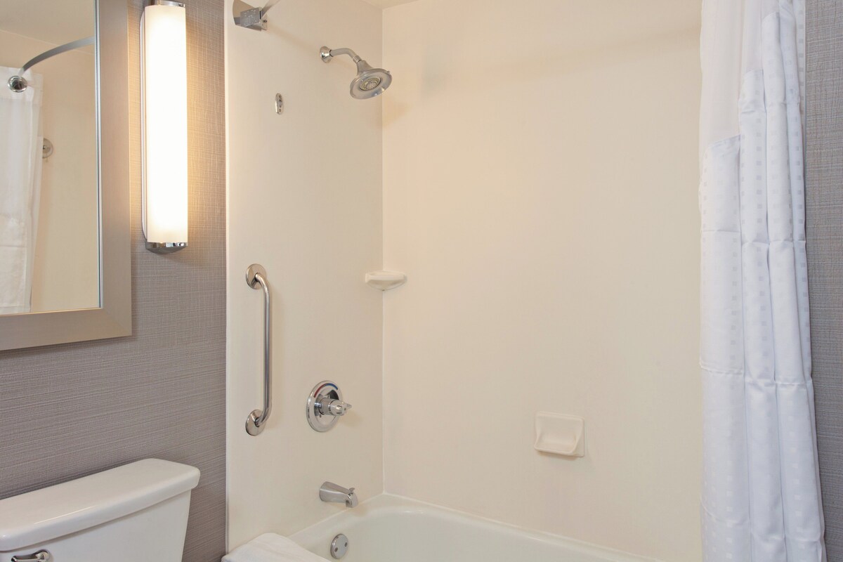 A bathroom with a toilet, a shower with a handheld showerhead, a grab bar, and a wall-mounted light fixture.