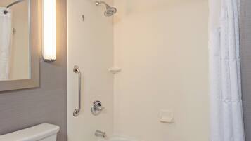 Un baño con inodoro, ducha con cabezal de ducha de mano, barra de apoyo y lámpara fijada a la pared.