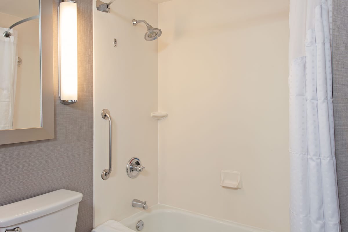 A bathroom with a toilet, a shower with a handheld showerhead, a grab bar, and a mirror with a light fixture.
