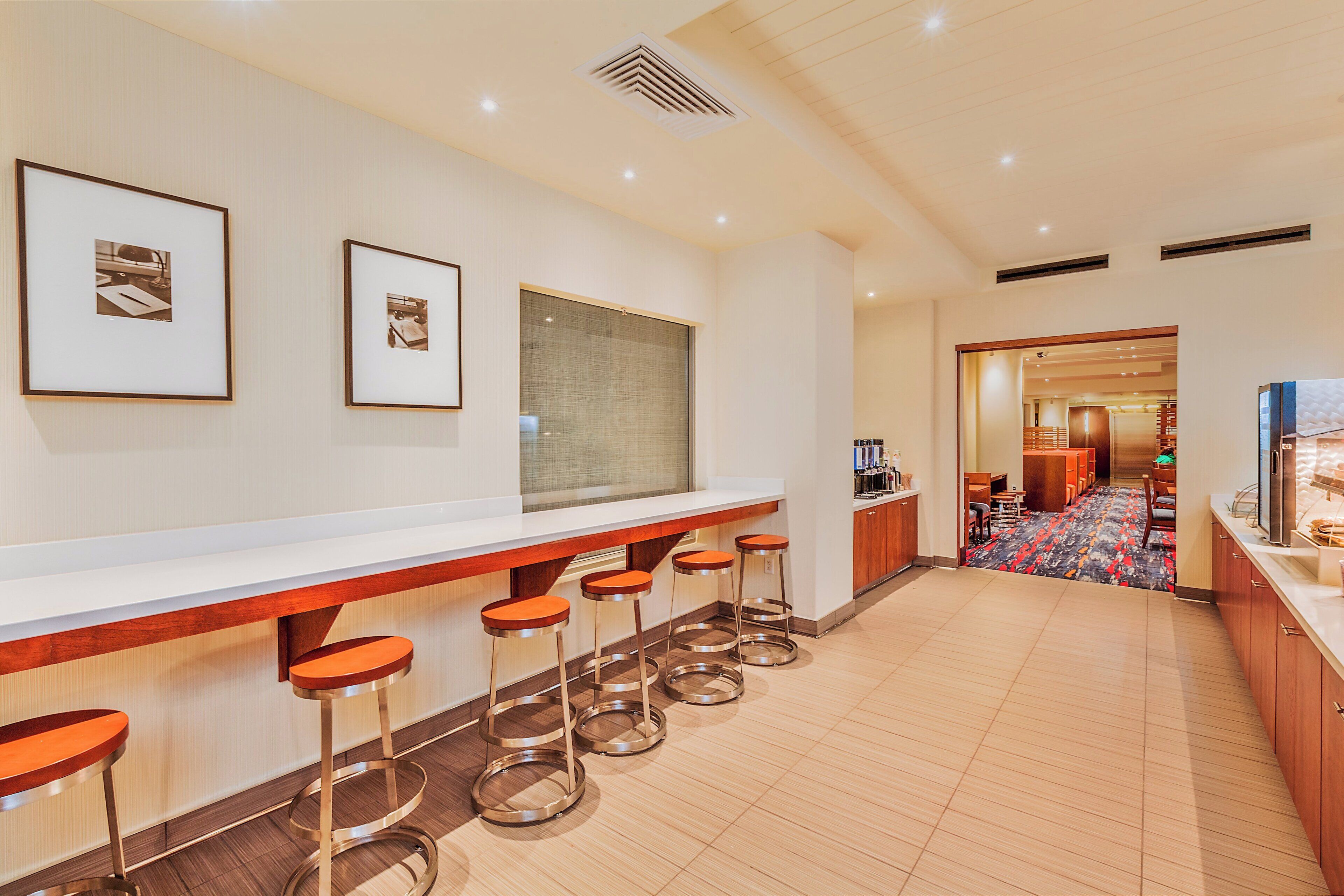 A hotel bar with a long counter, multiple stools, framed pictures on the wall, and a view into the dining area.