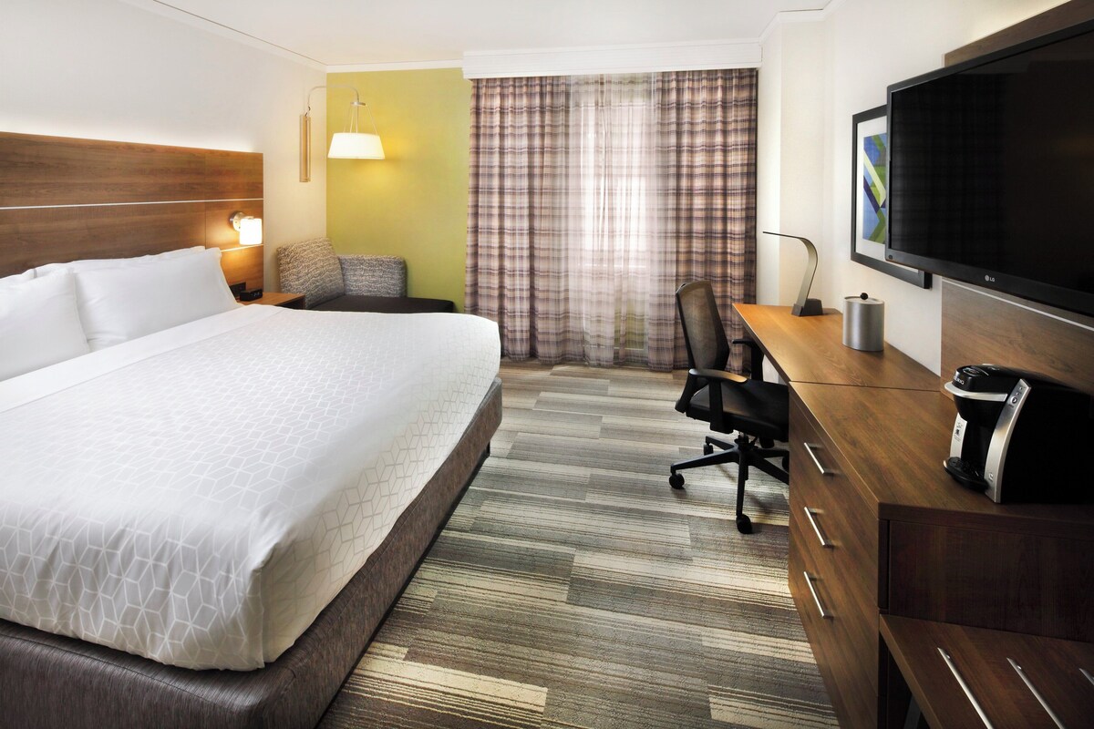 A hotel room with a large bed, a desk with a coffee maker, a TV, and a window with curtains.