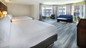 Une chambre d’hôtel avec deux lits, un canapé, une petite table et une vue sur les immeubles à travers de grandes fenêtres.