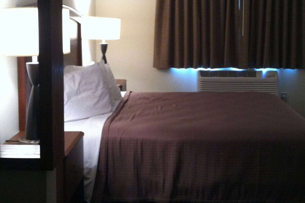 Una habitación de hotel con cama, mesita de noche, lámpara, ventana con cortinas y un radiador.