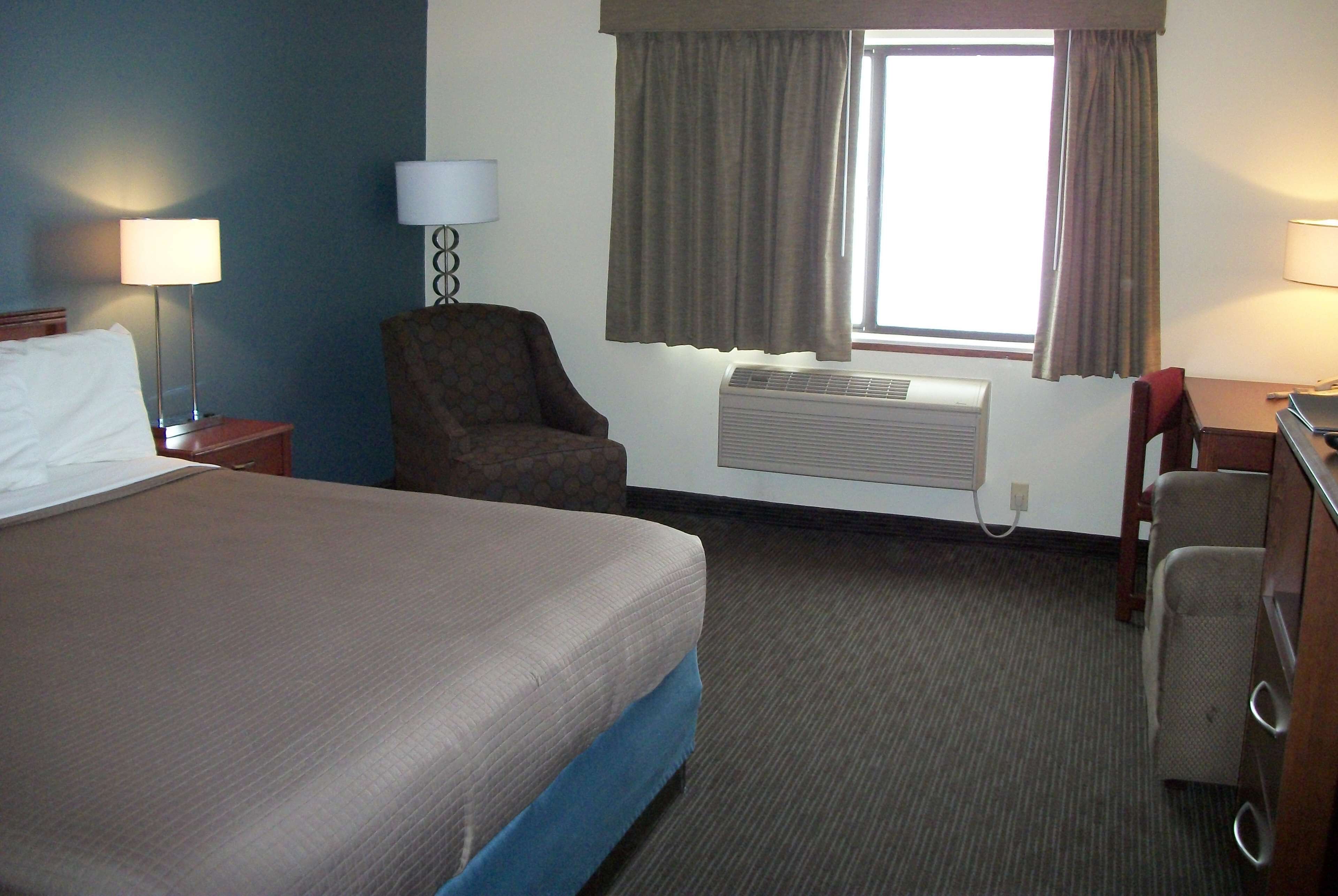 A hotel room with a bed, a chair, a desk, a lamp, and an air conditioning unit.