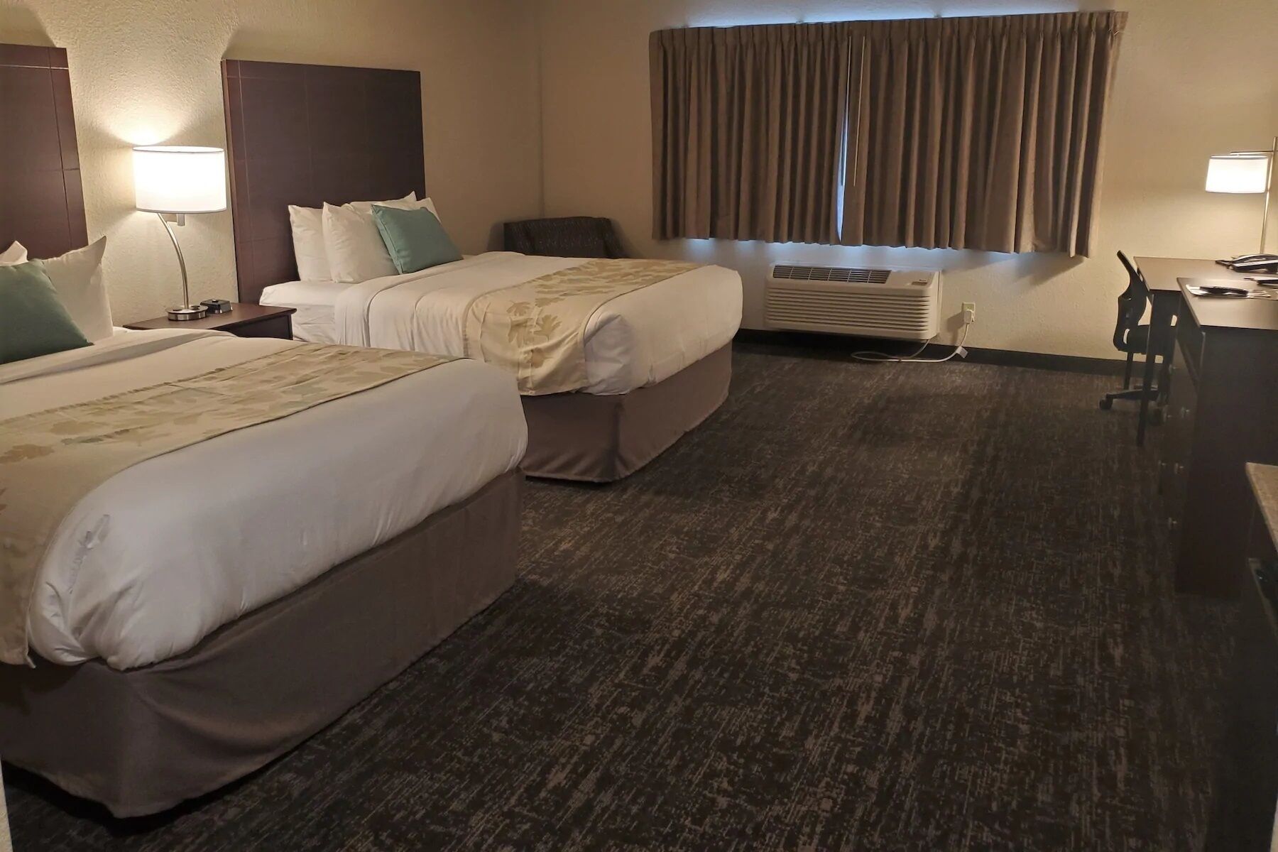 Habitación de hotel con dos camas, un escritorio, una silla, un equipo de aire acondicionado y una ventana con cortinas.