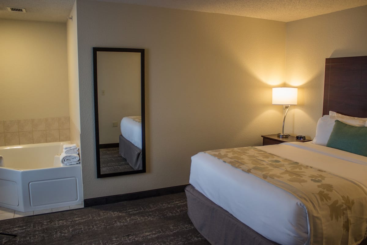 A hotel room with a bed, a nightstand with a lamp, a bathroom with a bathtub, and a mirror on the wall.