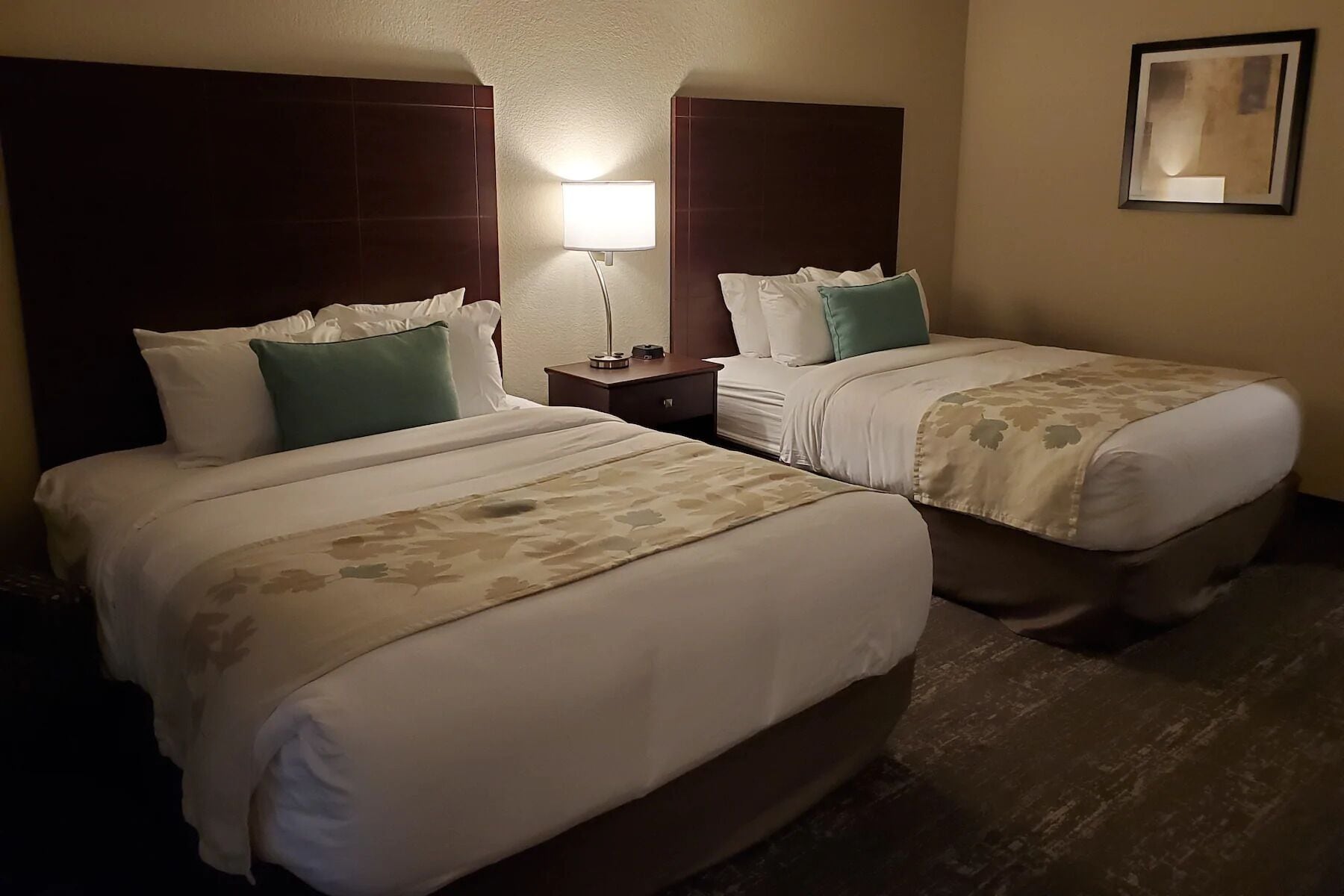 A hotel room with two beds, a nightstand, a lamp, and a framed picture on the wall.