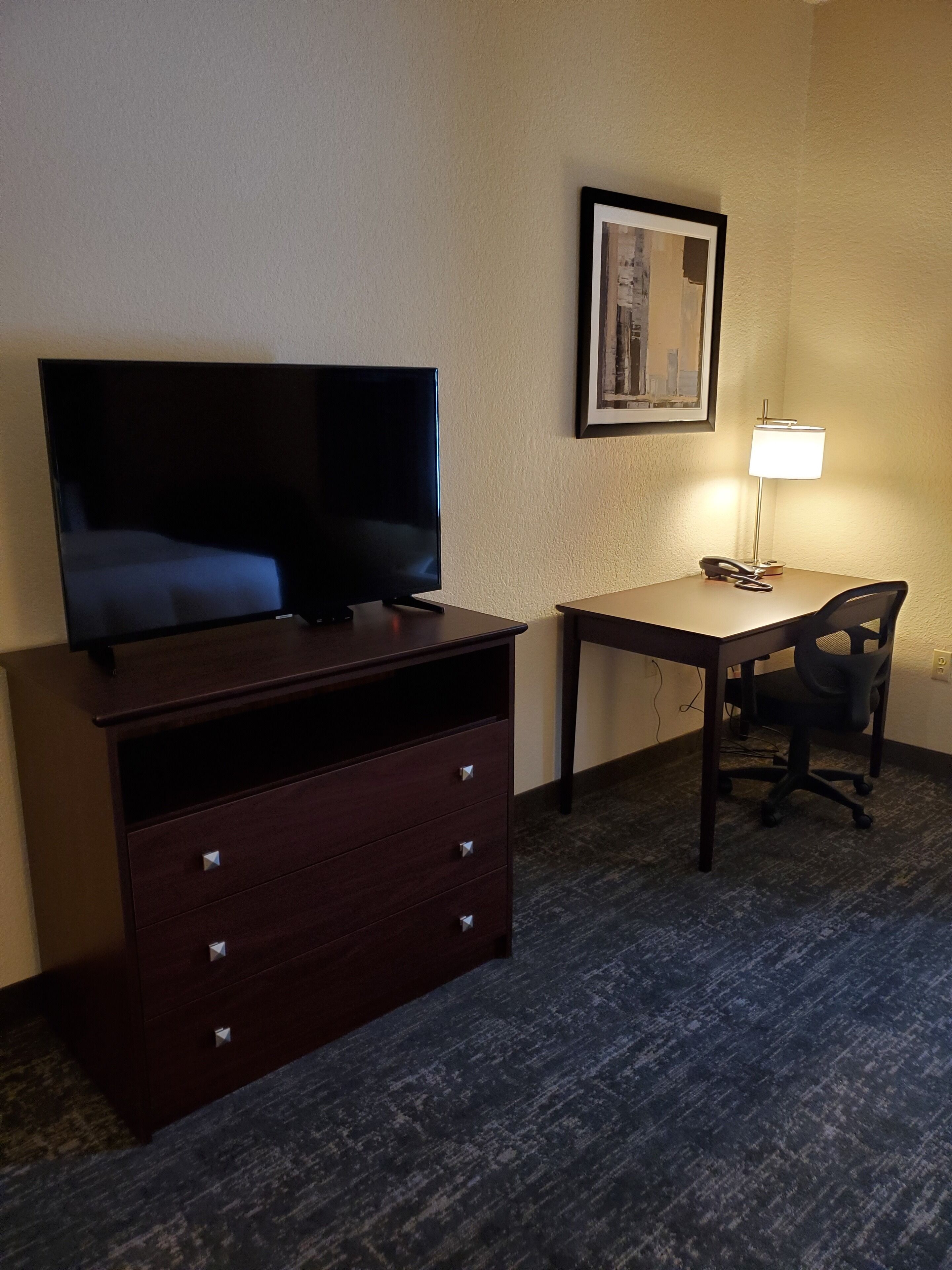 A hotel room with a flat-screen TV on a dresser, a desk with a chair, a lamp, and a framed picture on the wall.