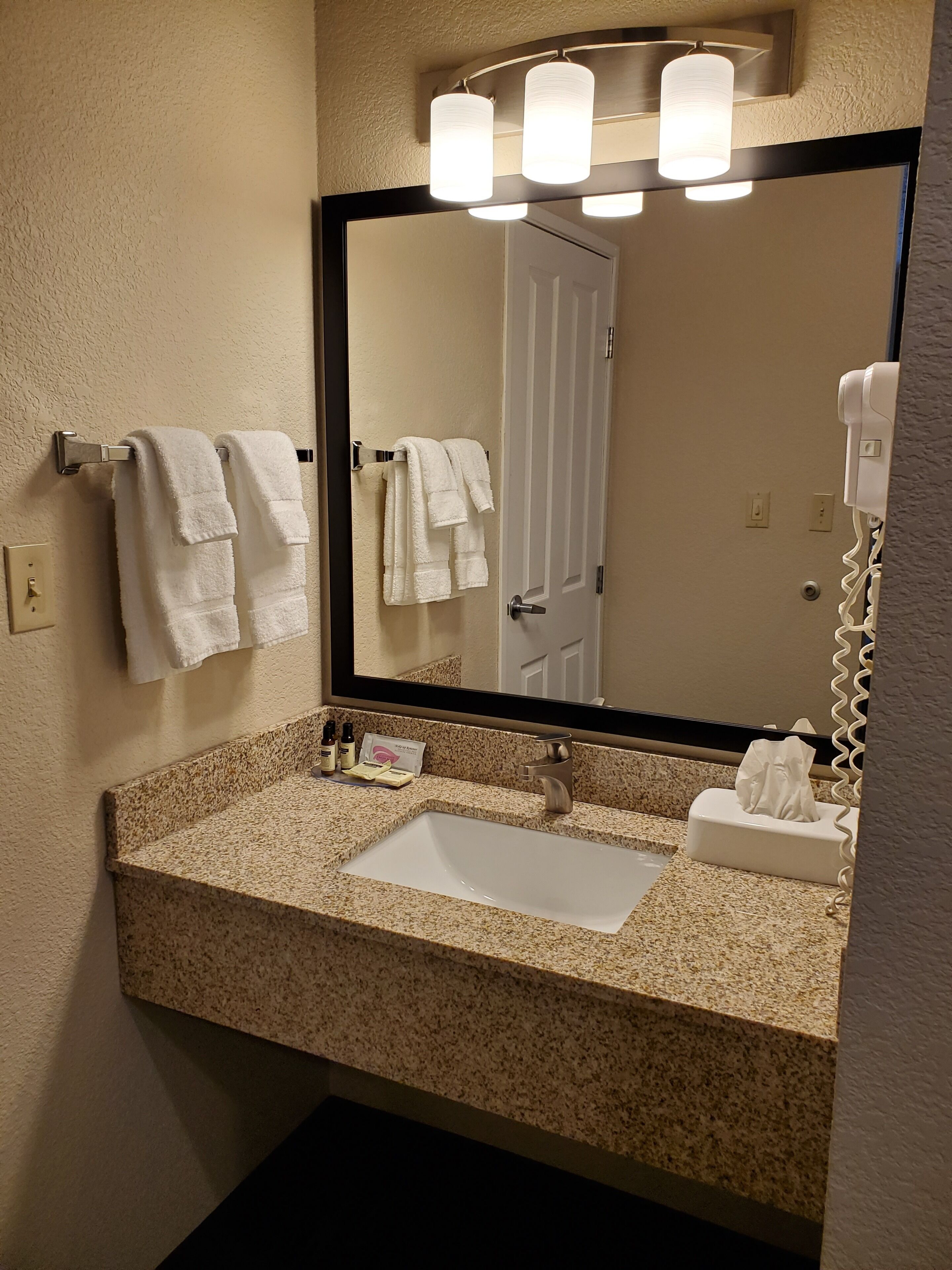A bathroom with a granite countertop sink, a mirror with three lights, and a towel rack with white towels.