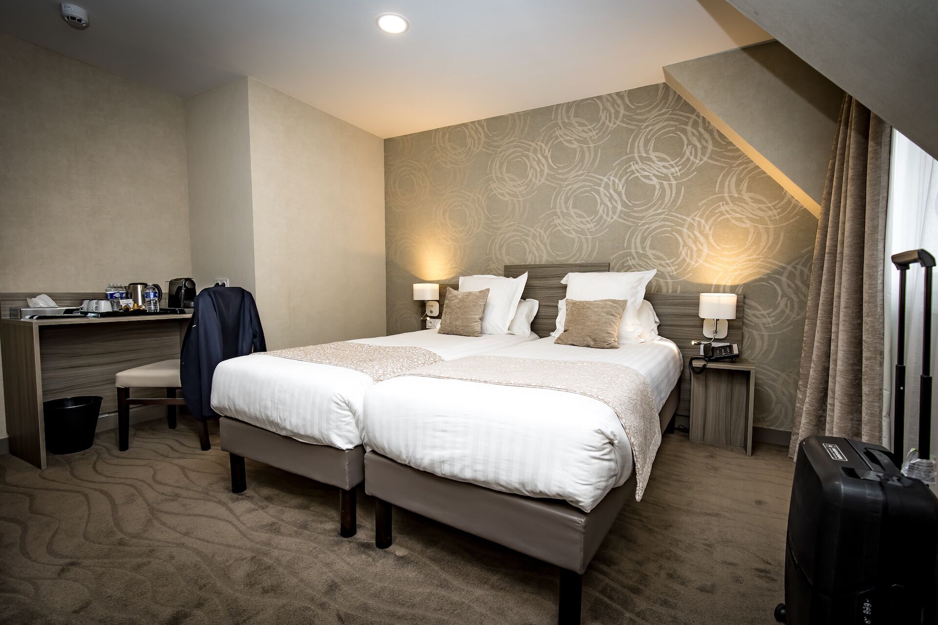 A hotel room with a large bed, a desk with a chair, a suitcase, and a wall-mounted lamp.
