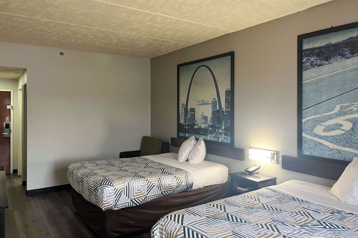 Hotellihuone, jossa on kaksi sänkyä, kaupunkimaisemaa esittävä taulu, yöpöytä lampun kanssa ja ovi vasemmalla puolella.