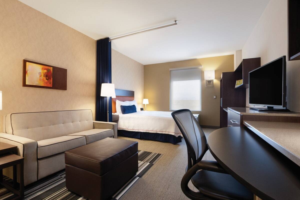 A hotel room with a sofa, a bed, a desk with a chair, a television, and a painting on the wall.