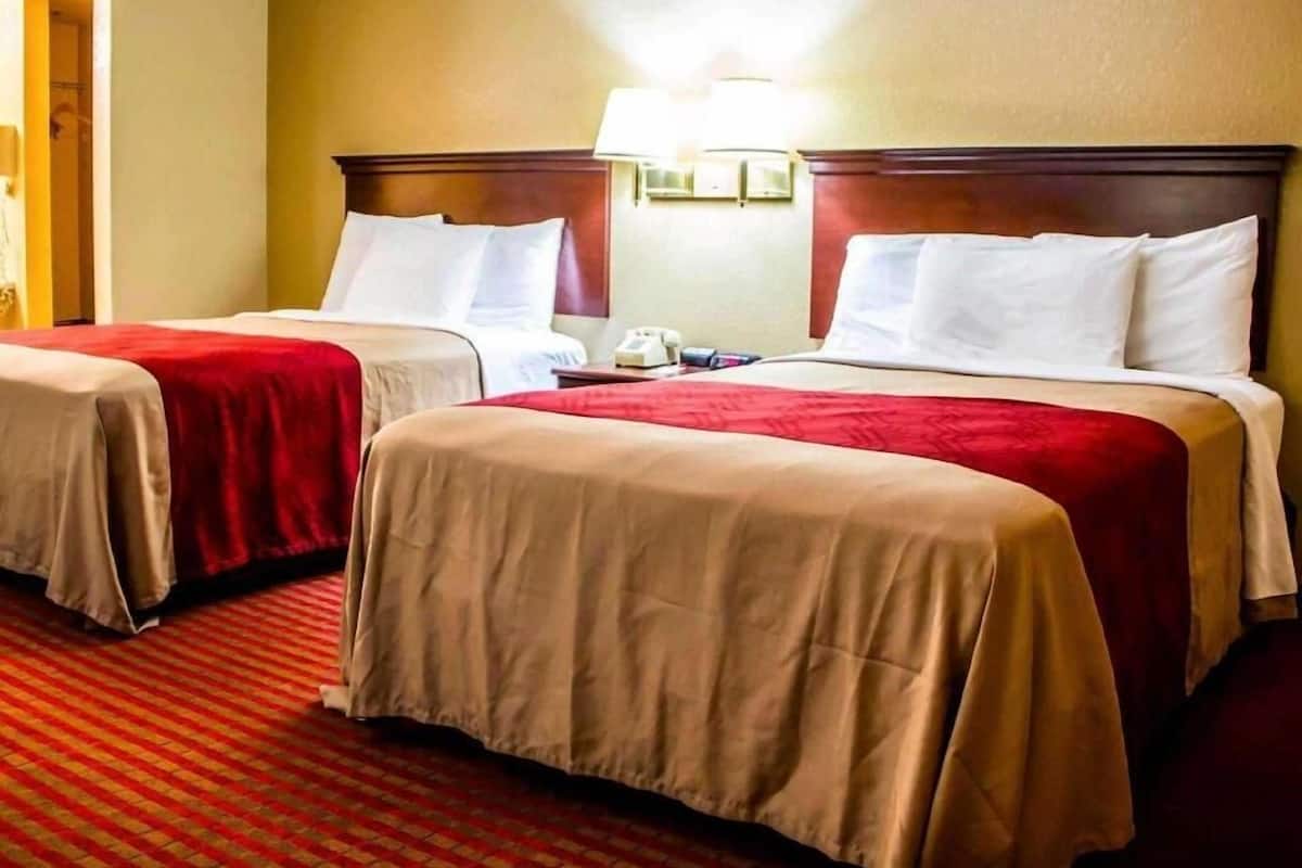Quarto de hotel com duas camas, cada uma com colcha vermelha e almofadas brancas, uma cabeceira de madeira, uma mesinha de cabeceira com abajur e piso coberto por carpete.