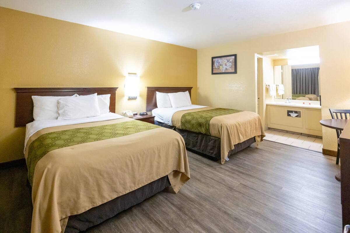 Quarto de hotel com duas camas, uma pequena mesa de trabalho, um espelho e um banheiro visível por uma porta aberta.