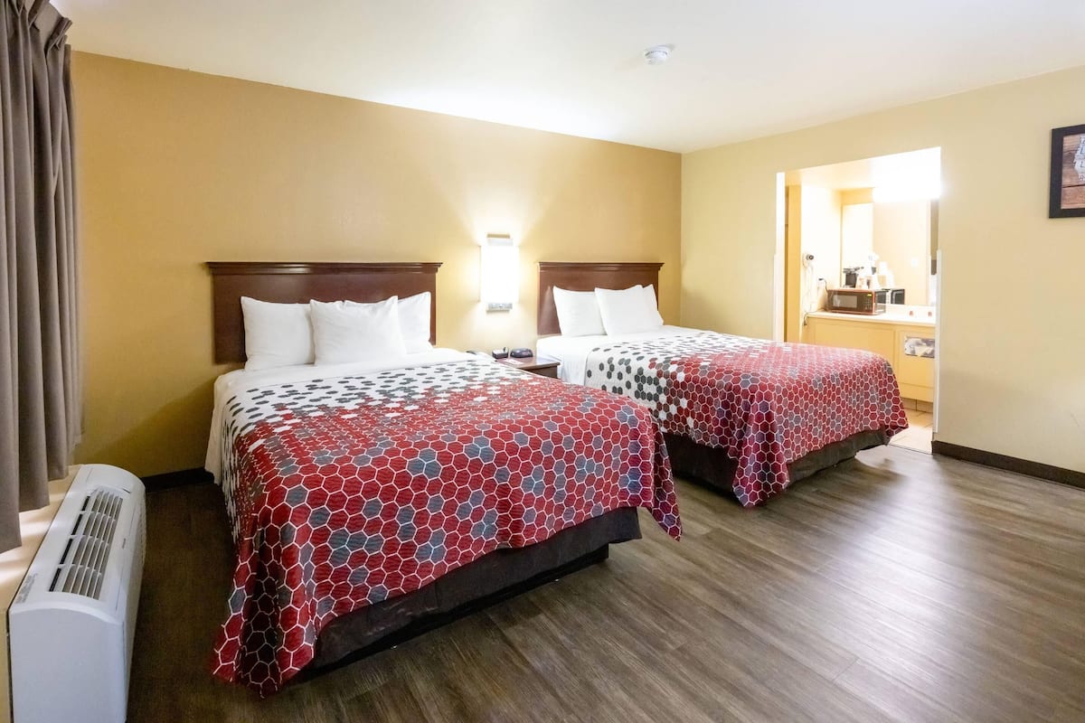 Quarto de hotel com duas camas, cada uma com cabeceira de madeira, colcha com padrão em vermelho e cinza, mesa de cabeceira com abajur e espelho fixado na parede.