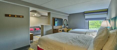 A hotel room with two beds, a desk, a mirror, and a window with a view of greenery.