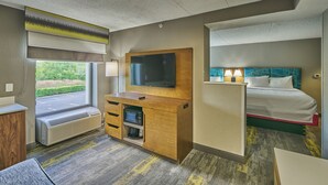 Habitación de hotel con cama, televisor, escritorio con computadora, microondas y ventana con vista a follaje.