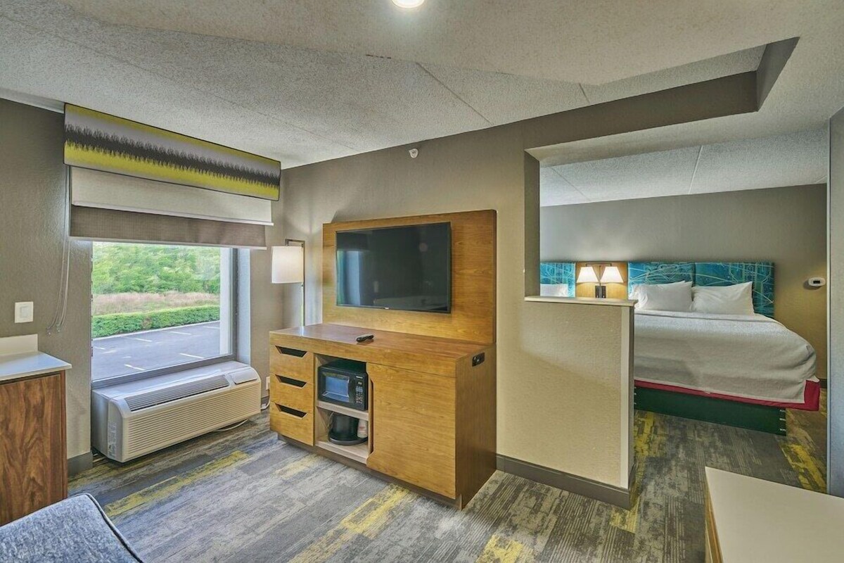 Habitación de hotel con cama, televisión, escritorio con computadora, cafetera y ventana con vistas al verdor.