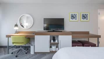 Habitación de hotel con una cama, un escritorio con silla, un televisor, un espejo y dos cuadros en la pared.