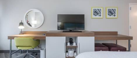 A hotel room with a bed, a desk with a chair, a television, a mirror, and two framed pictures on the wall.