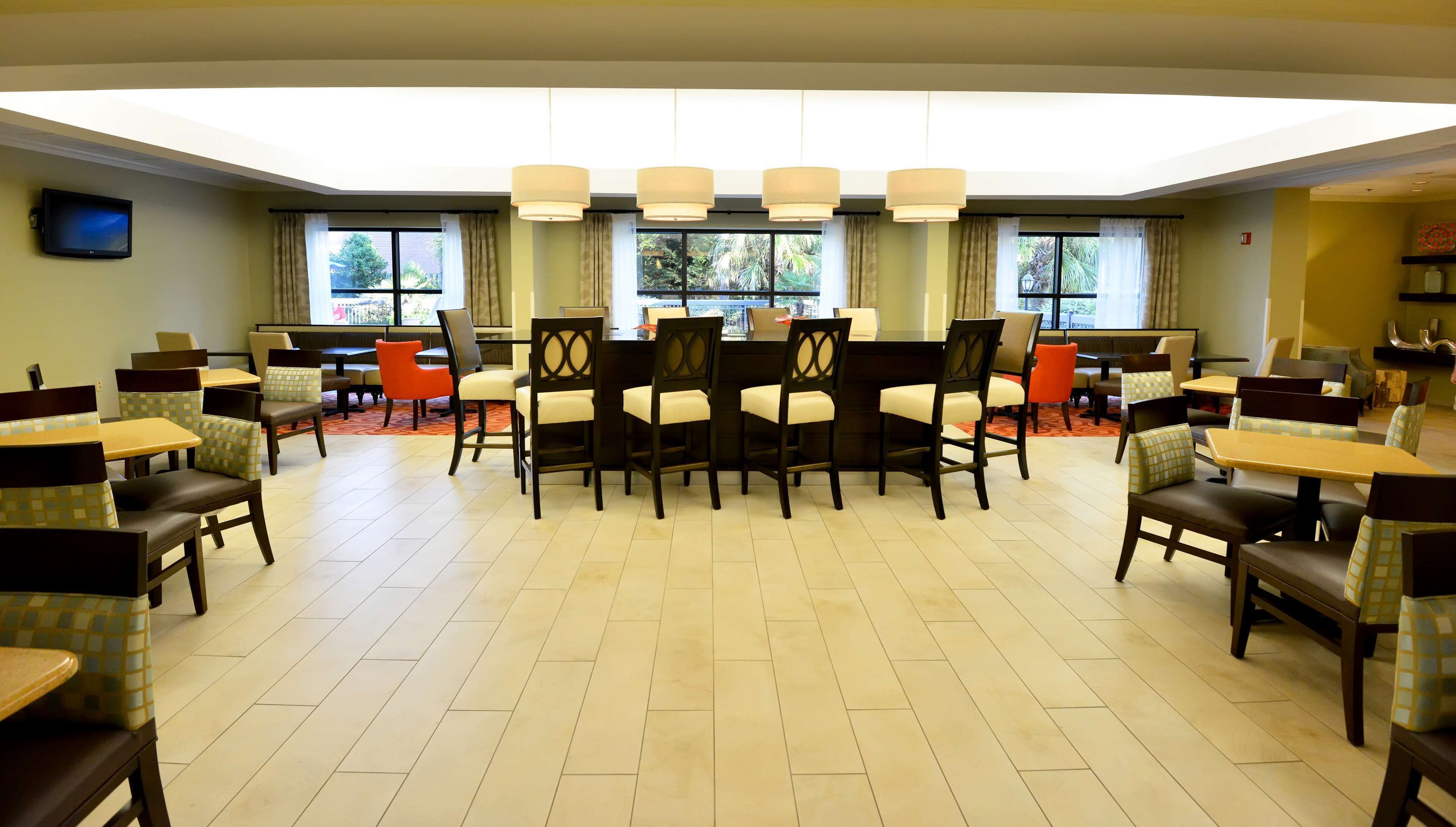 Un comedor de hotel con varias mesas y sillas, amplias ventanas con cortinas y un televisor de pantalla plana en la pared.