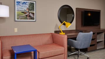 A hotel room with a red sofa, a blue side table, a grey office chair, a yellow desk lamp, a large mirror, and a flat-screen TV mounted on a wooden cabinet.