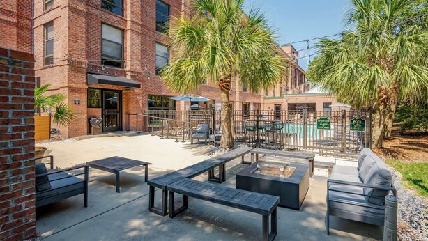 An outdoor patio with seating, palm trees, and a brick building with a pool area in the background. - Hampton Inn Wilmington-Medical Park (Wilmington)