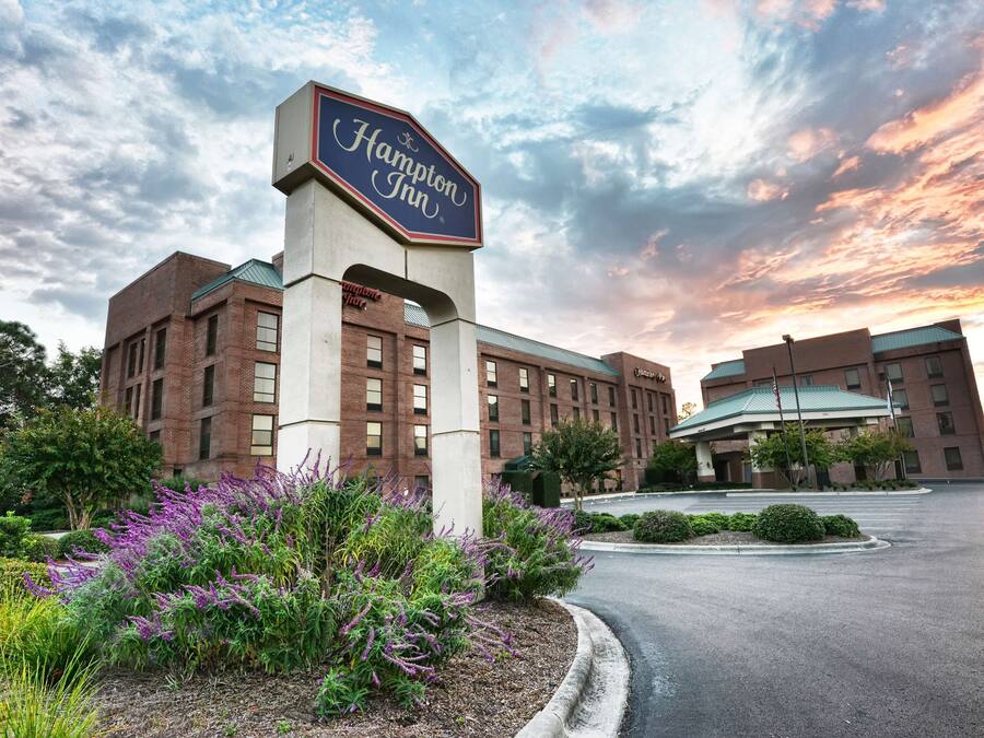 A Hampton Inn hotel with a prominent sign, surrounded by landscaped gardens and a parking lot.
