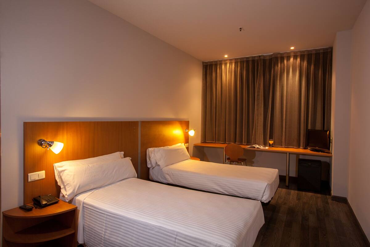 Habitación de hotel con dos camas individuales, un escritorio con silla, una mesita de noche con lámpara y un televisor.