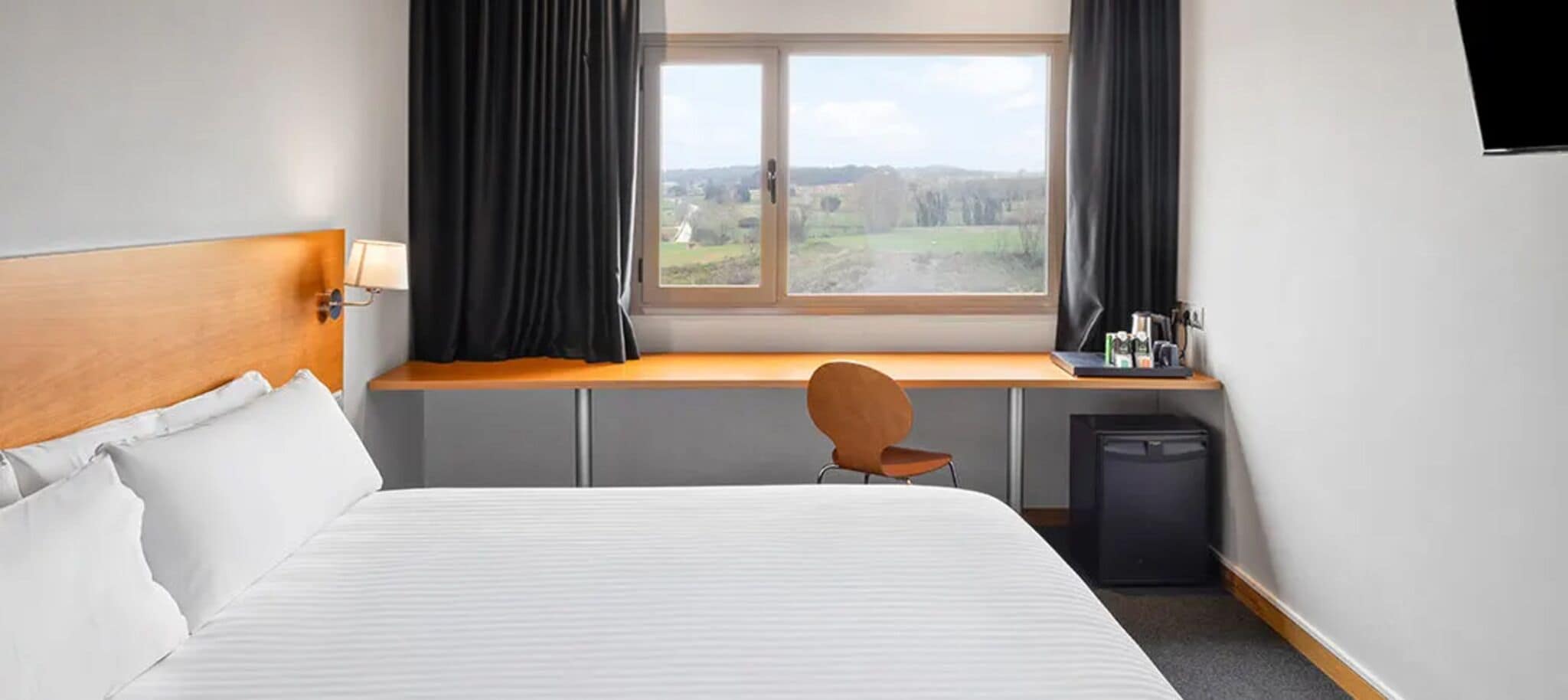 Une chambre d’hôtel équipée d’un lit, d’un bureau, d’une chaise, d’une fenêtre avec des rideaux et d’un petit réfrigérateur.