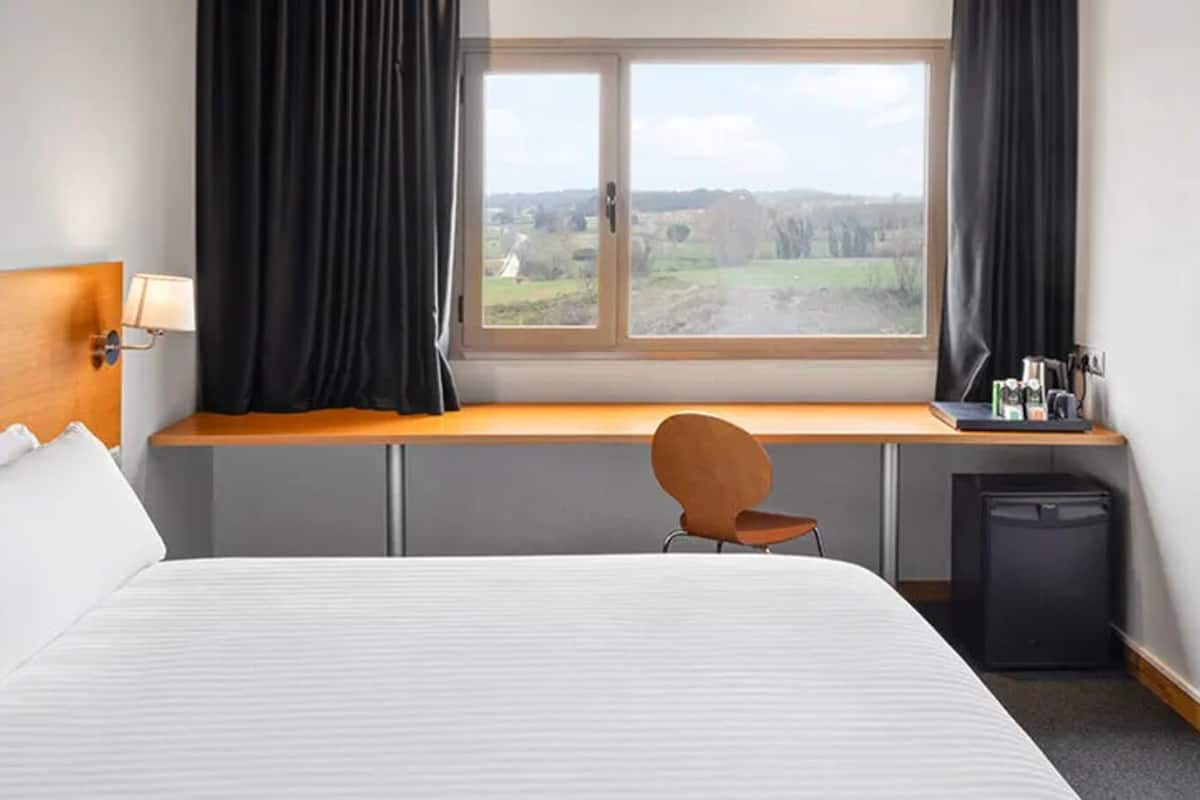 Une chambre d’hôtel équipée d’un lit, d’un bureau, d’une chaise, d’une fenêtre avec des rideaux et d’un petit réfrigérateur.