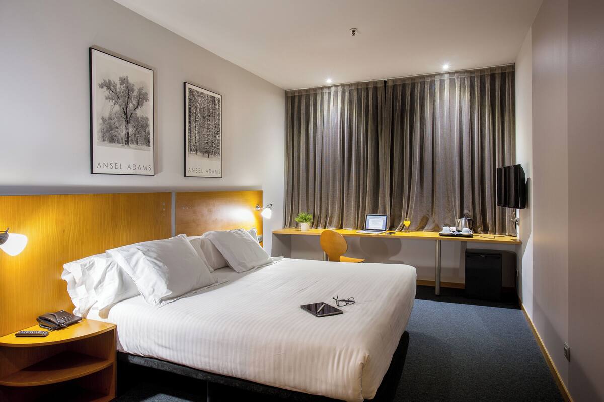 Une chambre d’hôtel comprenant un lit, une table de chevet, un bureau, une chaise et une fenêtre avec des rideaux.