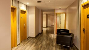 A hotel hallway with wooden doors, a checkered floor, and a seating area with chairs.