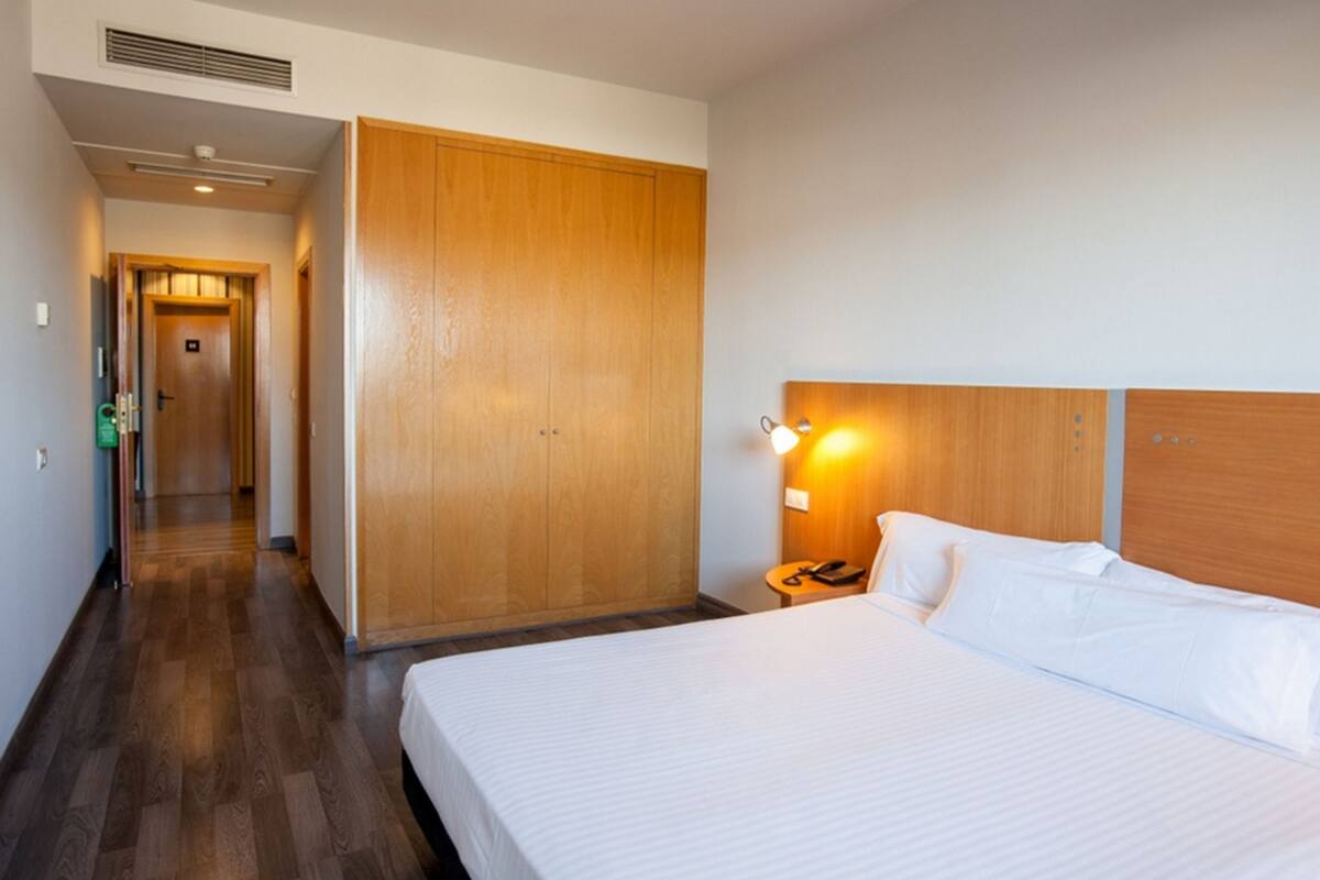 Habitación de hotel con un armario de madera, cama con ropa de cama blanca, mesita de noche con lámpara y un pasillo que conduce a otra habitación.