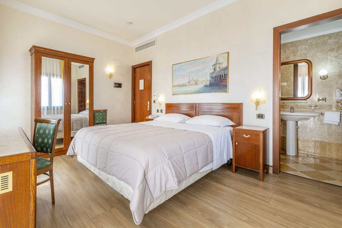 A hotel room with a bed, bedside tables, a desk, a chair, a mirror, and a painting on the wall.