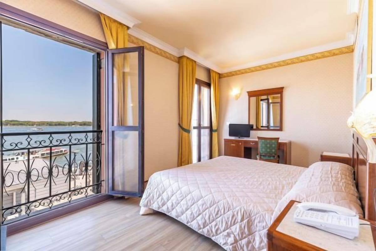 A hotel room with a bed, a desk, a chair, a TV, and a balcony with a view of the water.