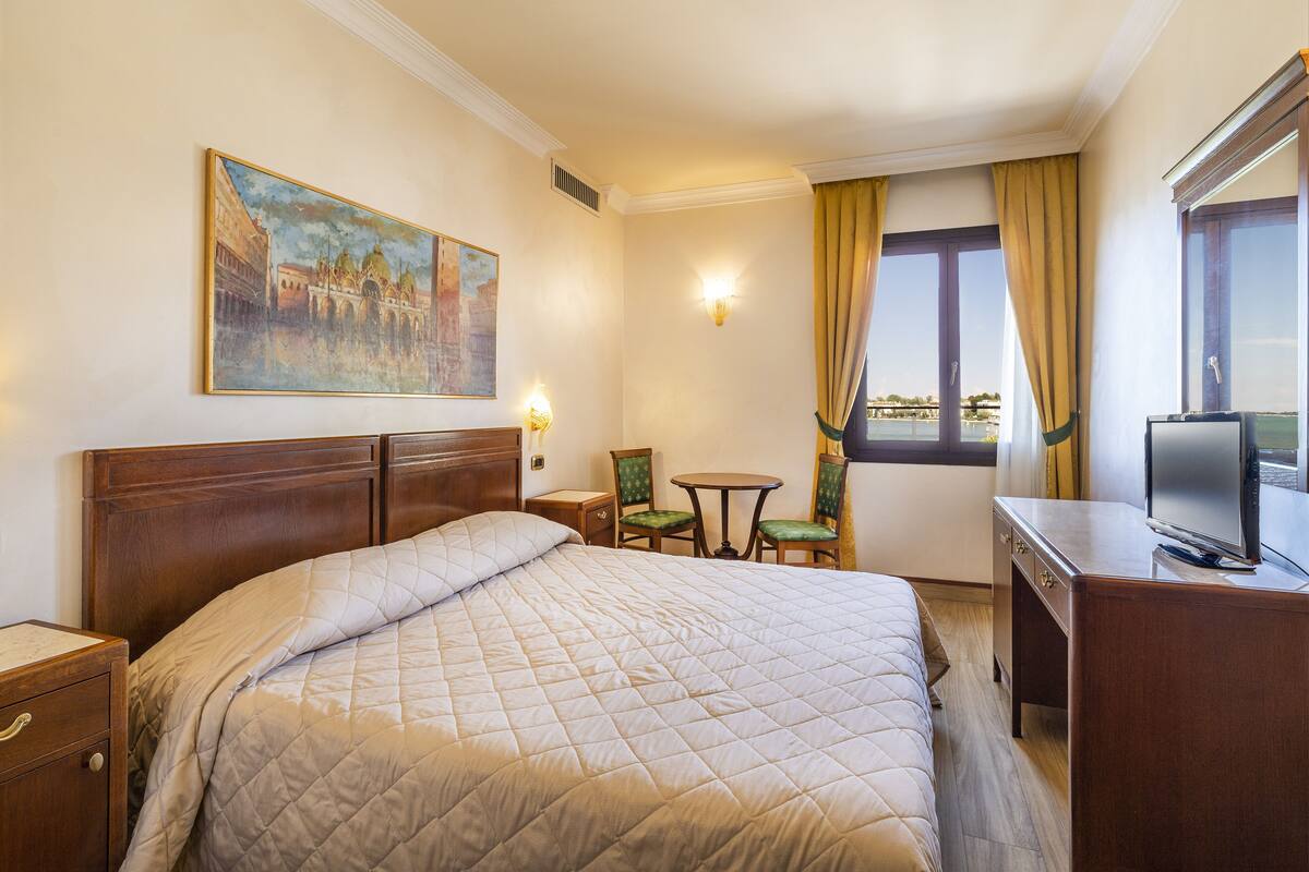 A hotel room with a large bed, a desk with a television, a small table with chairs, and a painting on the wall.